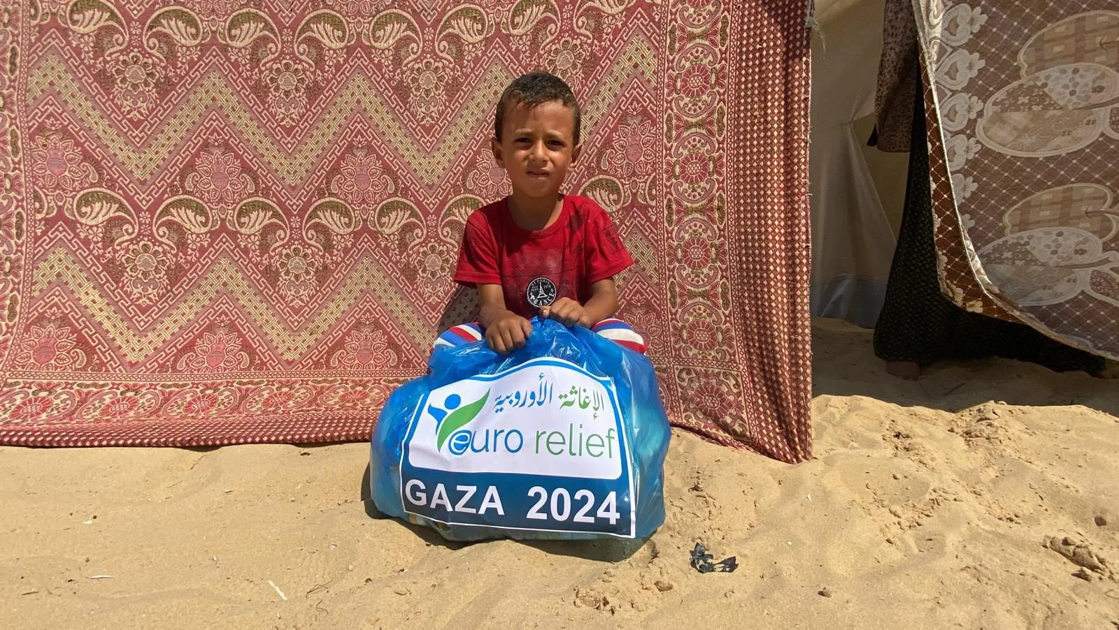 A boy holds an aid parcel beside his family tent